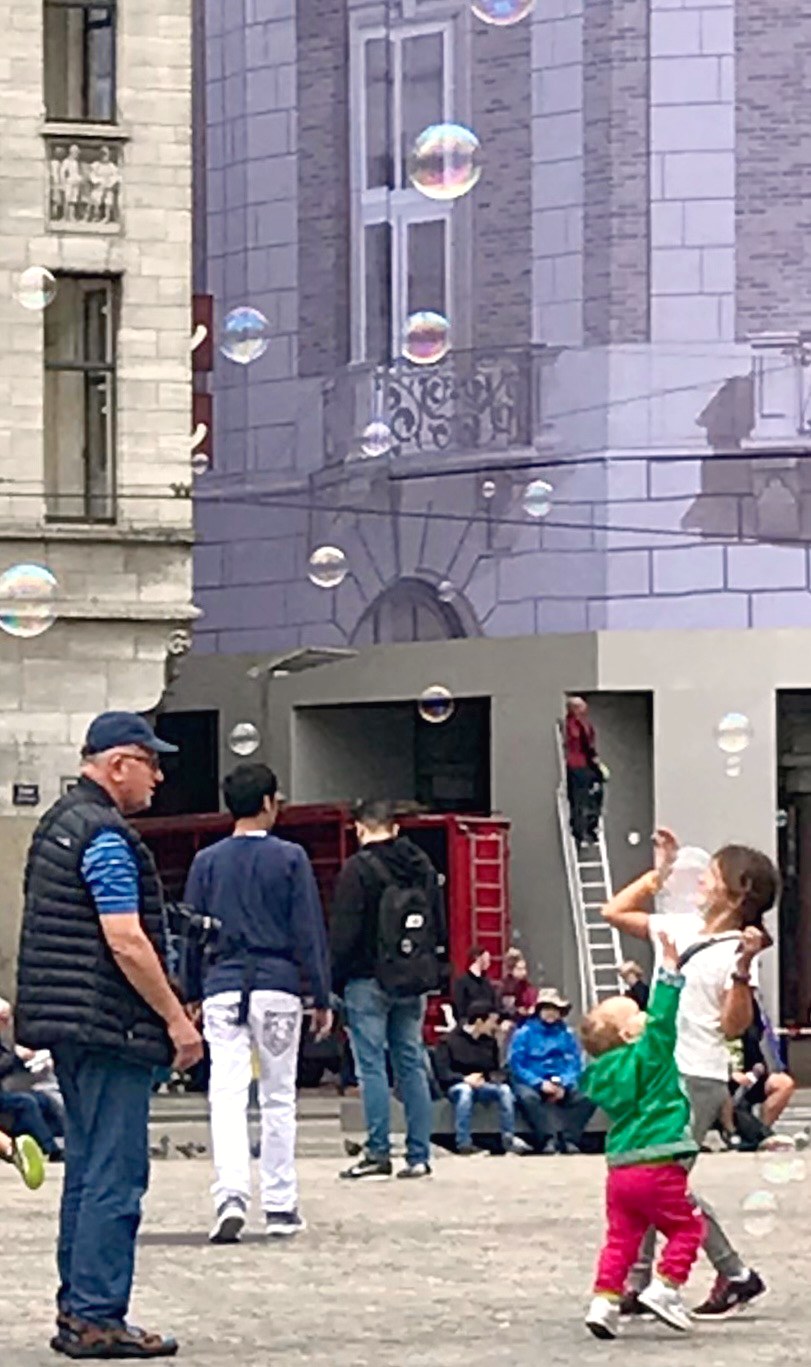 A child in a green jacket reaching for giant soap bubbles on Dam Square, watched by the bubble performer and passers-by