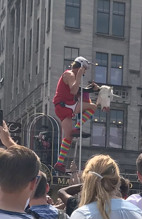 A street performer in rainbow socks and a seagull hat perched high on a pole with a unicorn puppet, above the Dam Square crowd